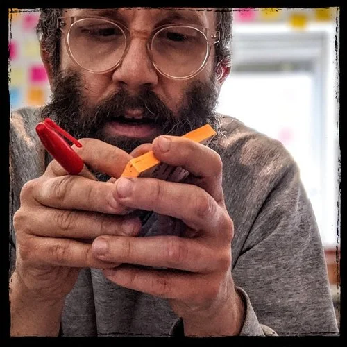 A man with glasses and a gray shirt holding a small orange object with a red clip, focused on it.