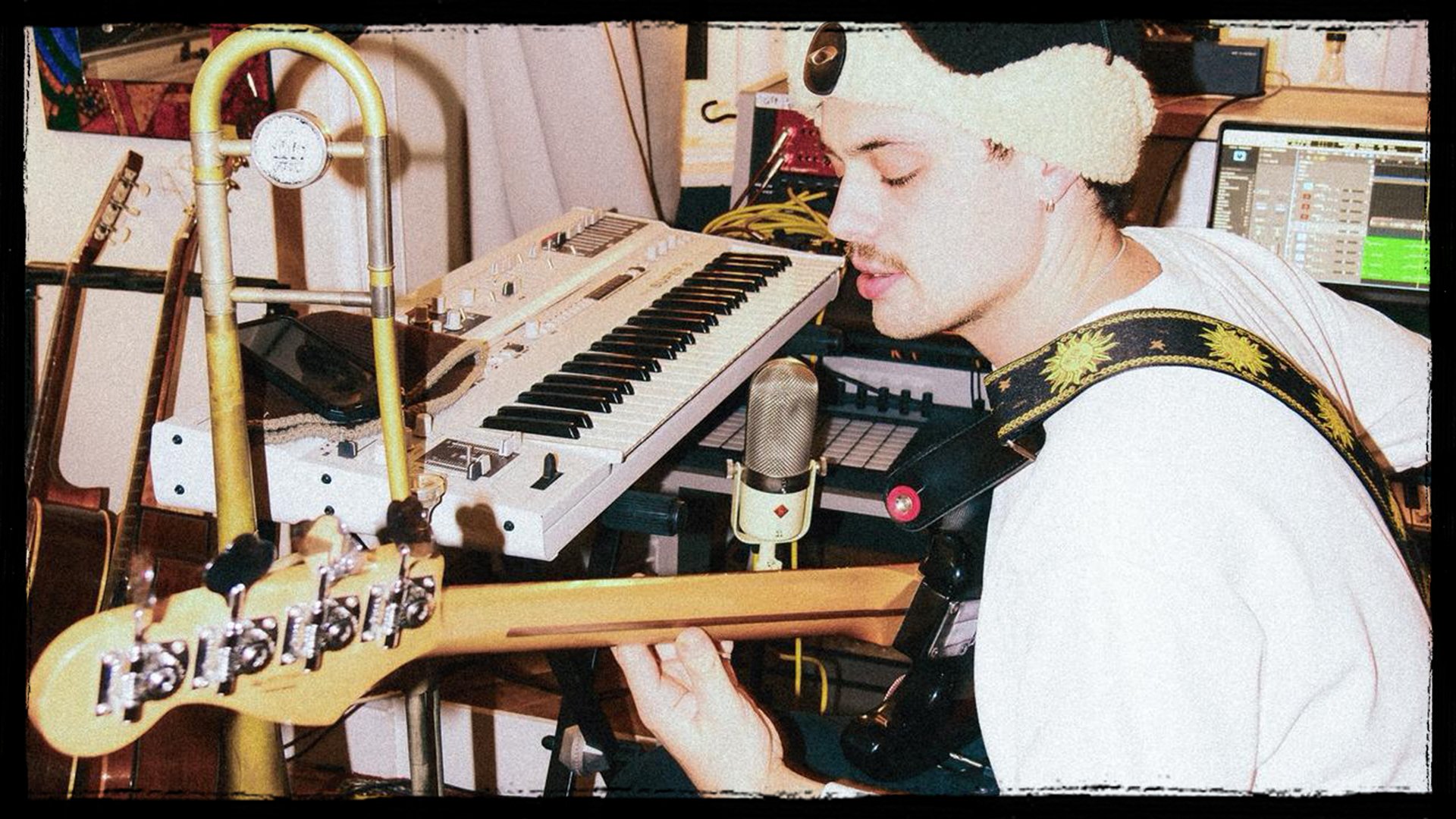 A musician with closed eyes singing into a microphone while playing an electric guitar, with a keyboard and computer in the background.