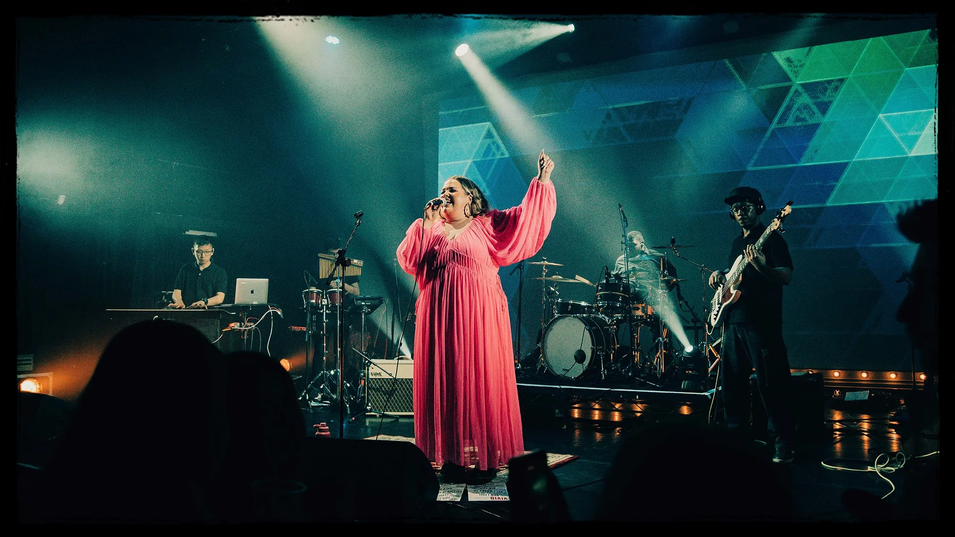 A singer in a bright pink dress performs with a band on stage, with a drummer, guitarist, keyboard player, and visual background projection.