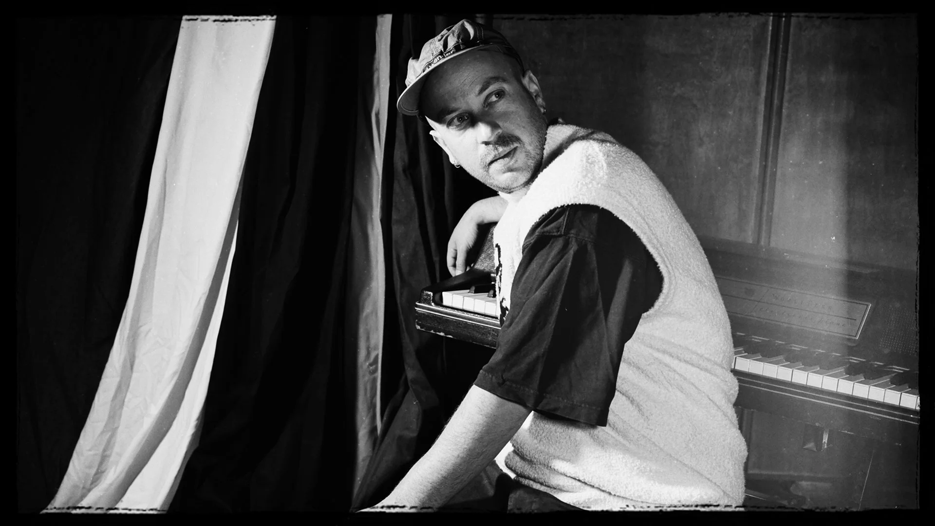 Black and white photo of a man sitting next to an upright piano, looking to the side with a serious expression, wearing a baseball cap and a vest over a dark shirt.
