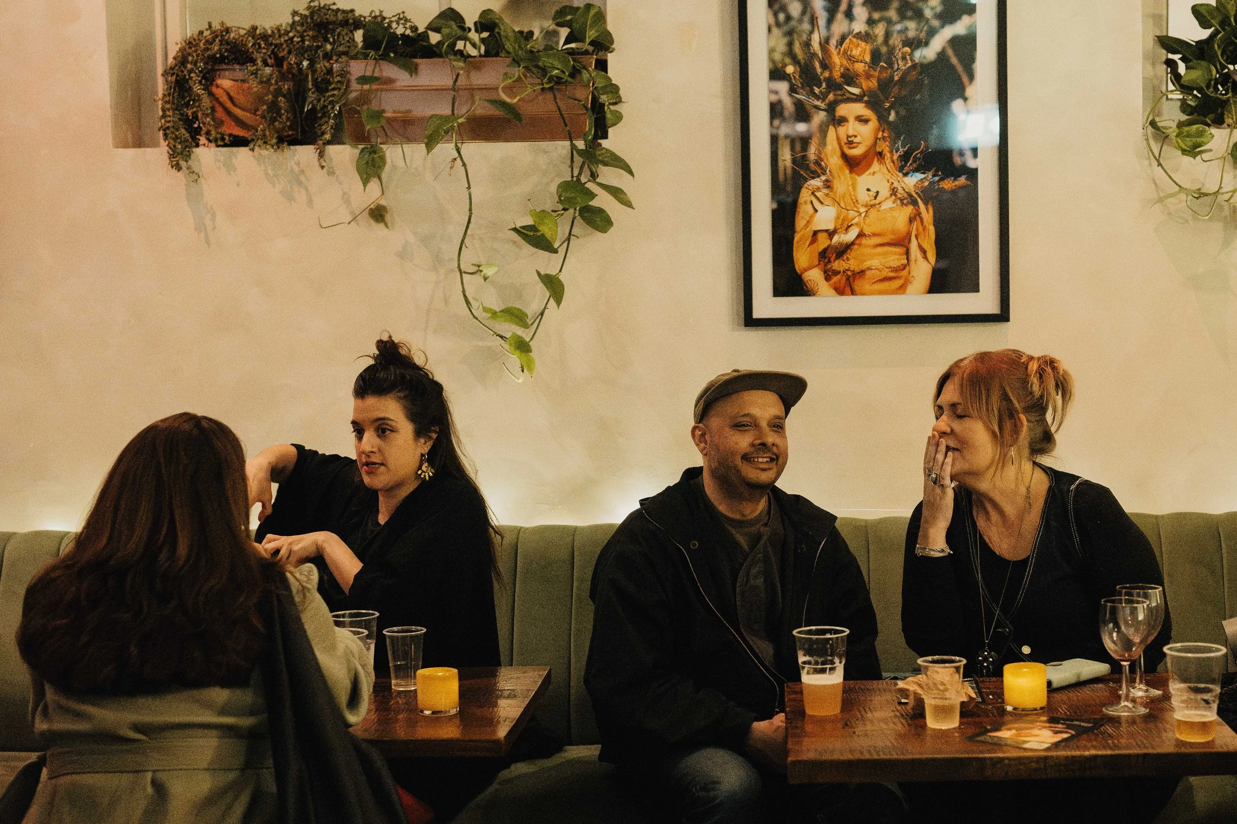Group of four people sitting at a restaurant table, engaged in conversation. Two women and two men are visible, with drinks on the table. The background shows a framed art piece of a woman with leaves in her hair, and hanging plants on the wall.