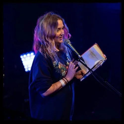 A woman with wavy blonde hair reading from a book at a microphone on stage under blue and purple lighting.