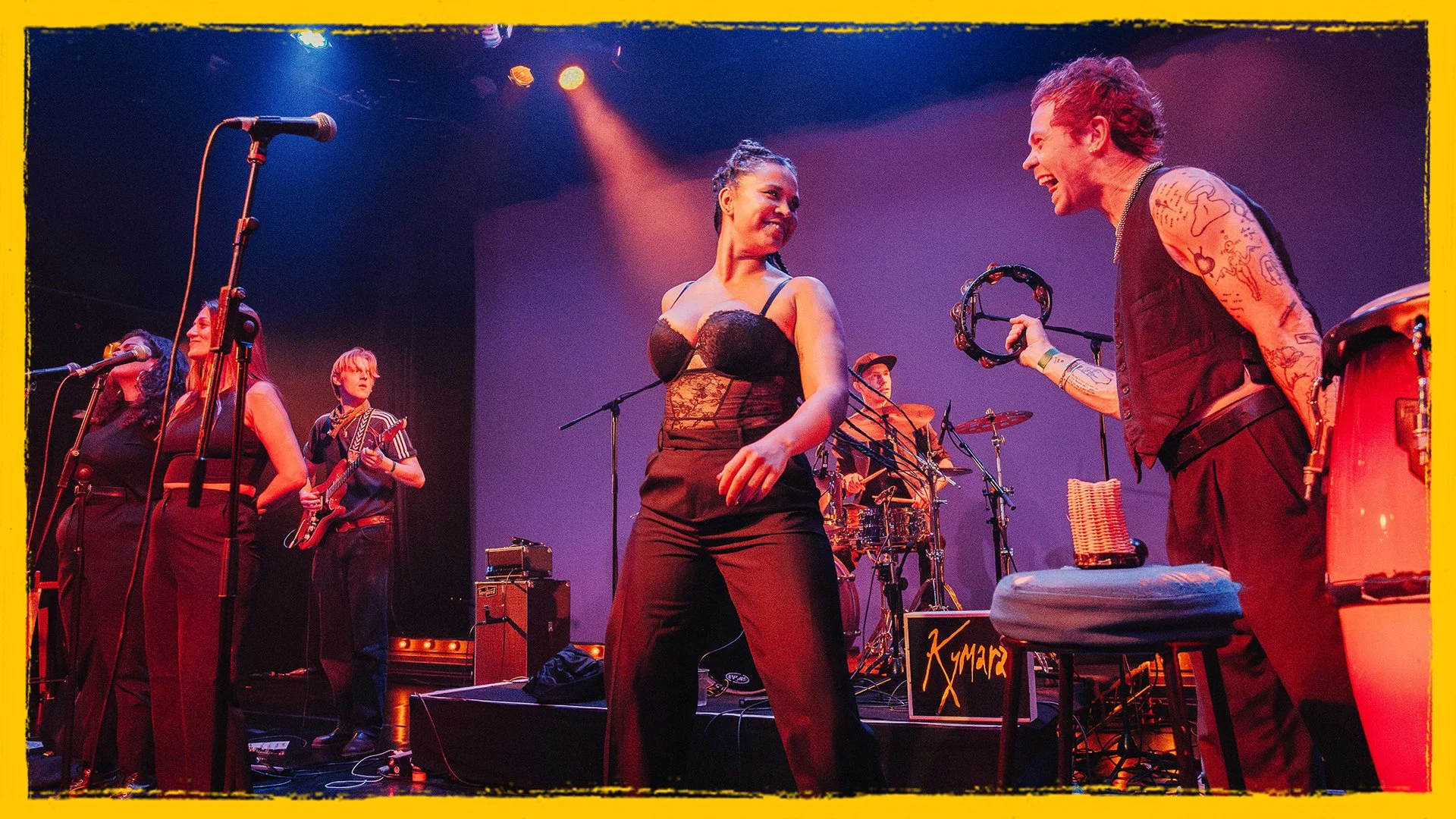 Musicians performing on stage, including a woman in black lace top and pants dancing with a man holding a tambourine, with other band members playing instruments in the background, under colorful stage lights.