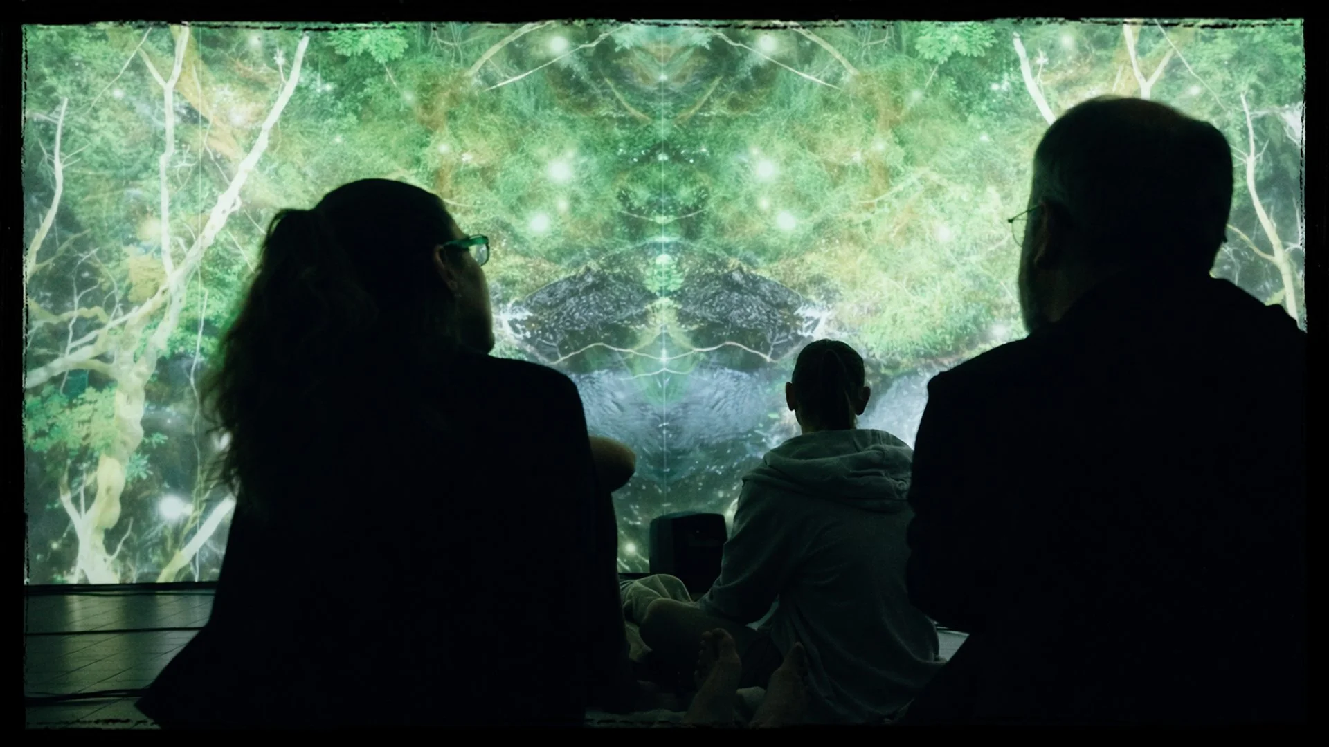 A family of three sitting on the floor in front of a large digital screen showing a vibrant, green forest scene with trees and glowing lights, creating an immersive experience.