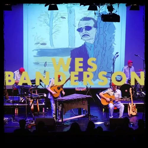 Music band performing on stage with large yellow letter signs spelling 'WES BANDERSON' in front of a screen showing a black and white illustration of a man with glasses and a mustache, trees, and falling leaves.