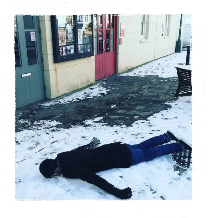 Person lying face down on snowy sidewalk outside building with colorful doors and windows.