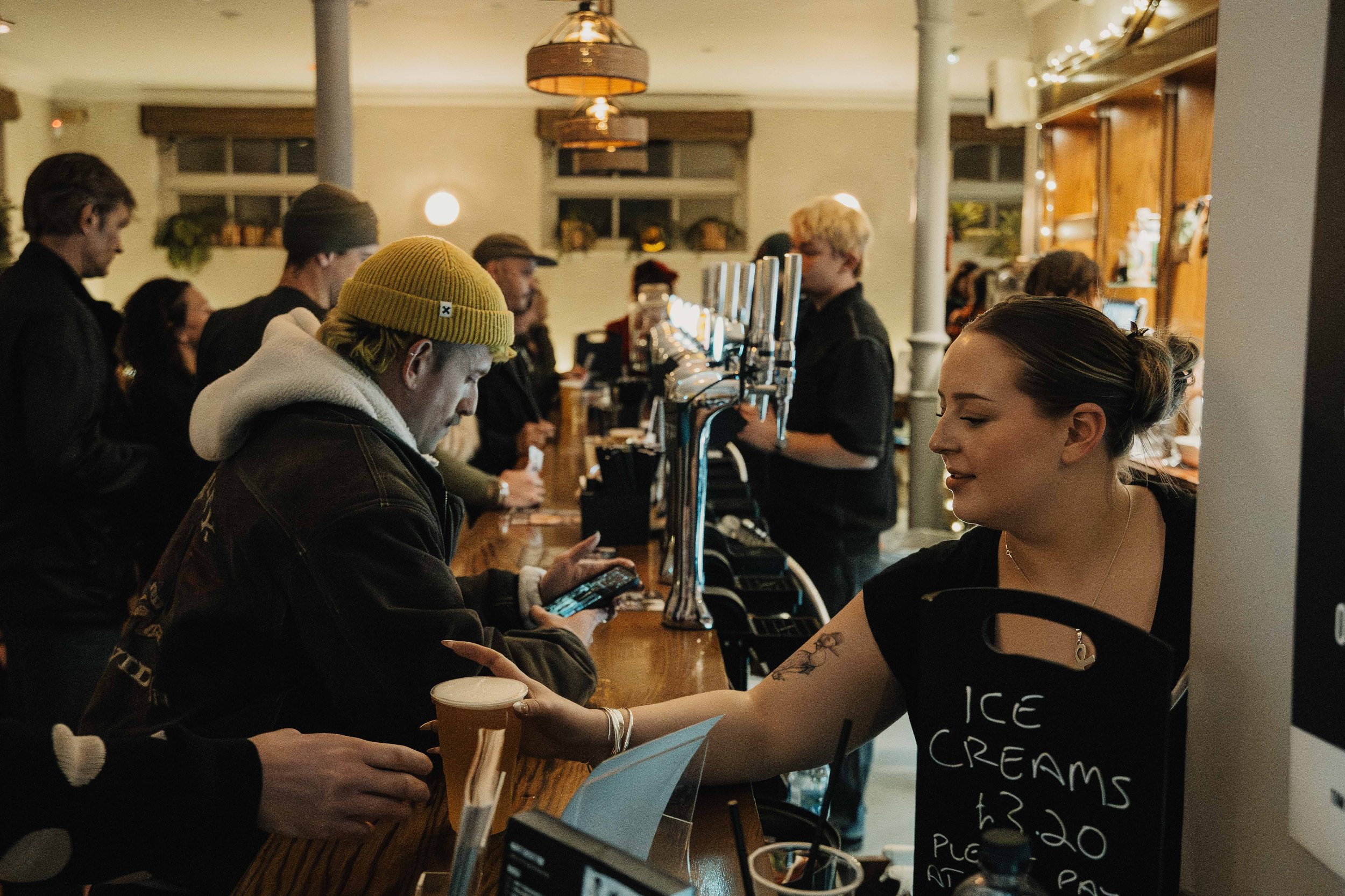 A woman at a bar handing a drink to a customer while several other patrons are in line or seated inside a cozy pub with warm lighting and wooden decor.