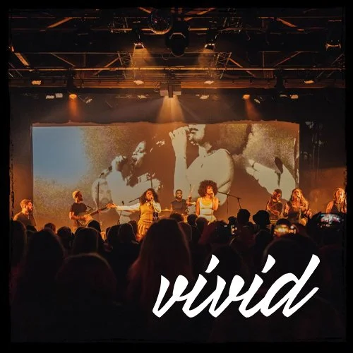 Live music concert with a band performing on stage, audience in the foreground, large screen behind band displaying black and white images, warm stage lighting, and the word "vivid" in white script overlay.