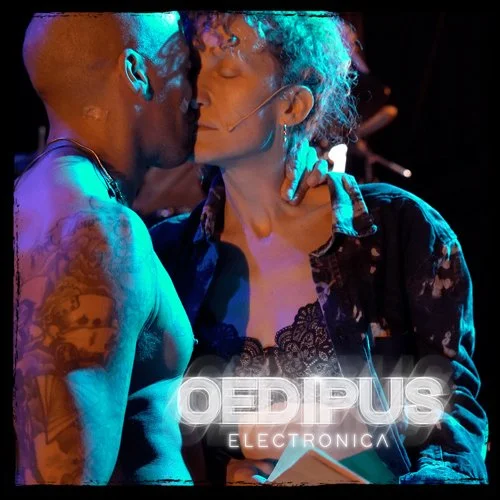 A man and woman, both with tattoos, sharing a close moment on stage during a performance, with the title 'Oedipus Electronica' displayed below them.