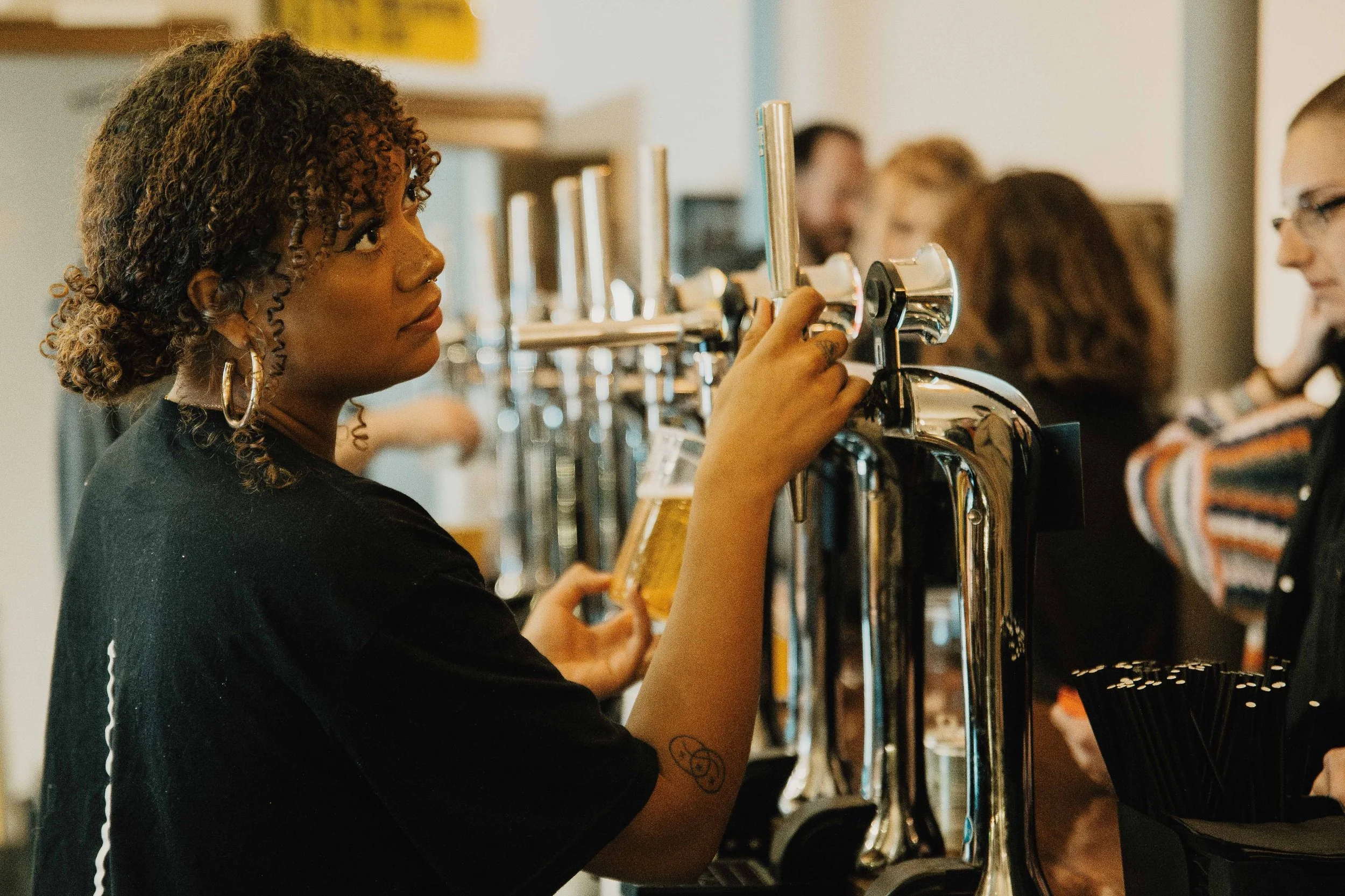 A woman with curly hair and hoop earrings pours a beer at a bar, surrounded by other patrons.