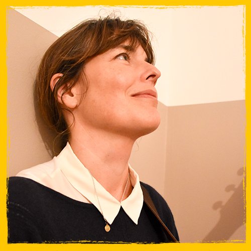 A woman with short brown hair wearing a white collared shirt and black sweater, looking upward with a gentle smile, standing against a beige wall.