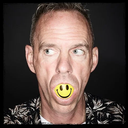 A man with short hair and a patterned shirt holds a yellow smiley face mask in his mouth, making a humorous expression.