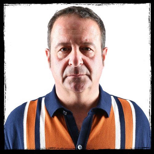 A middle-aged man with short dark hair, wearing a collared shirt with vertical orange, navy, white, and beige stripes, posing against a white background.