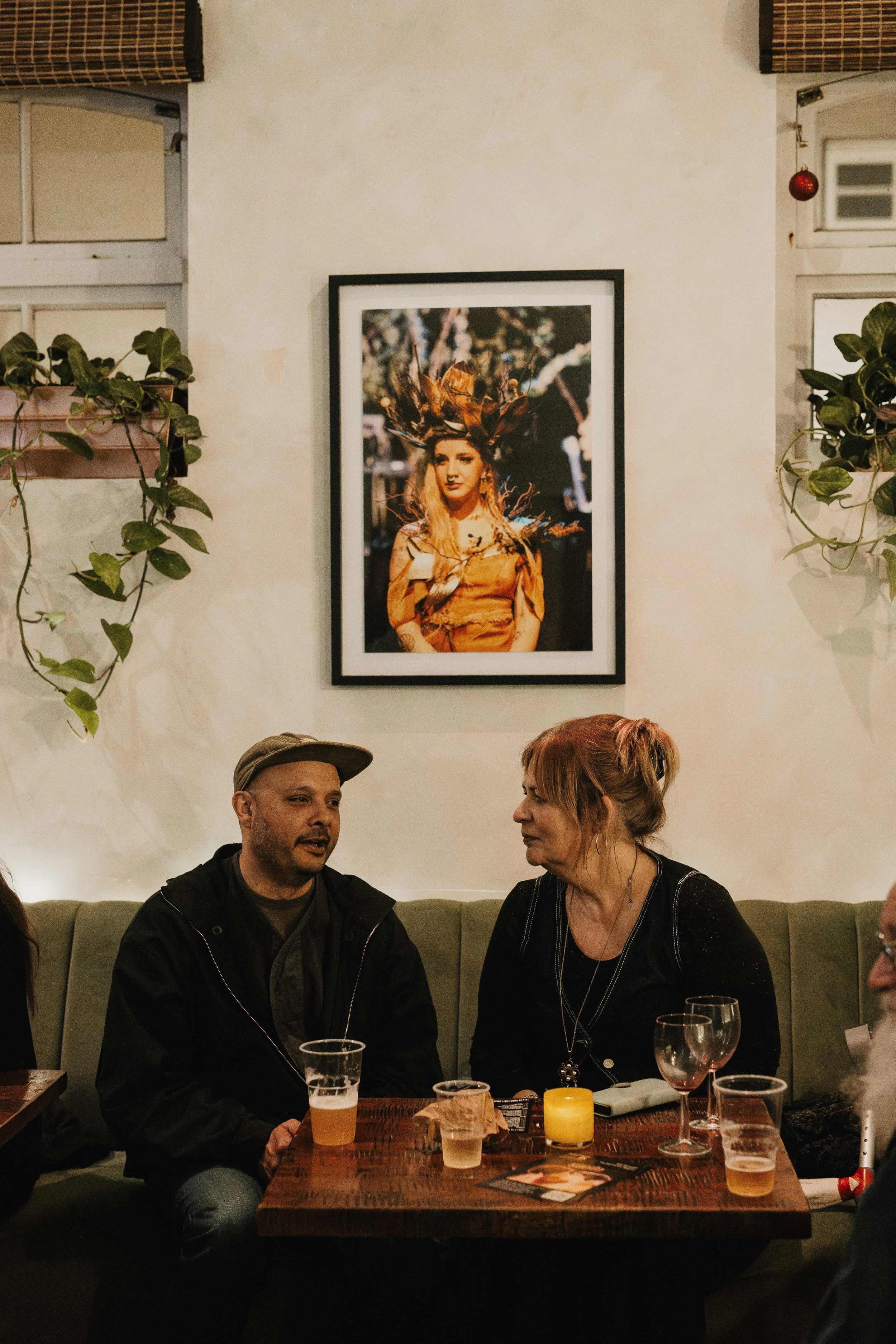 Two people sit at a table in a restaurant, engaged in conversation. Behind them, a framed photograph of a woman in an elaborate outfit hangs on the wall. The table has drinks and lit candles, and potted plants decorate the space.