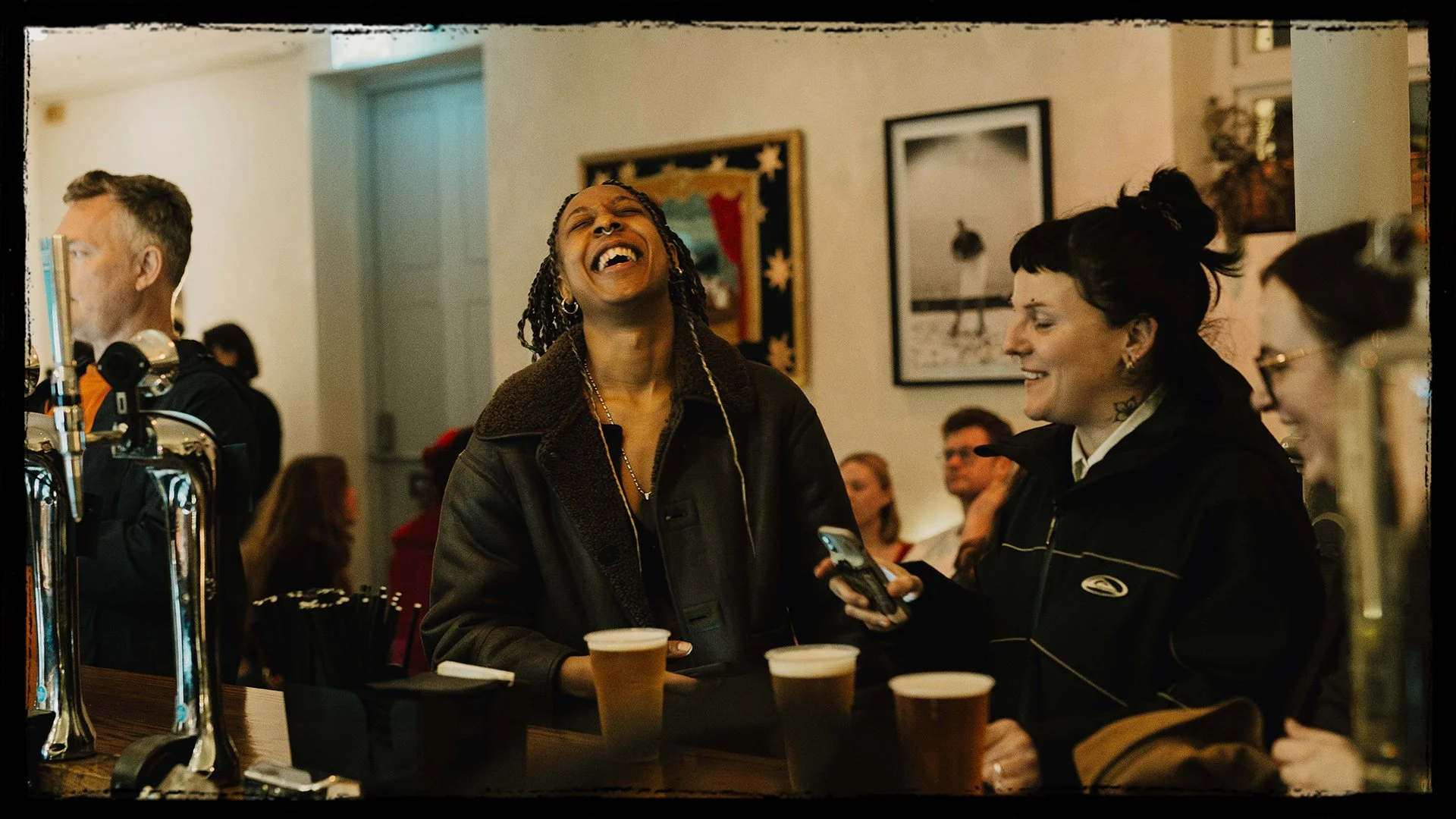 Group of people socializing at a bar, with a woman in the center laughing and holding a smartphone.