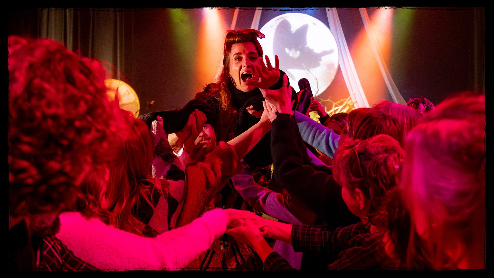 A woman with red hair and a bow in her hair in front of a full moon and spooky backdrop, being lifted by a crowd at a Halloween-themed event.