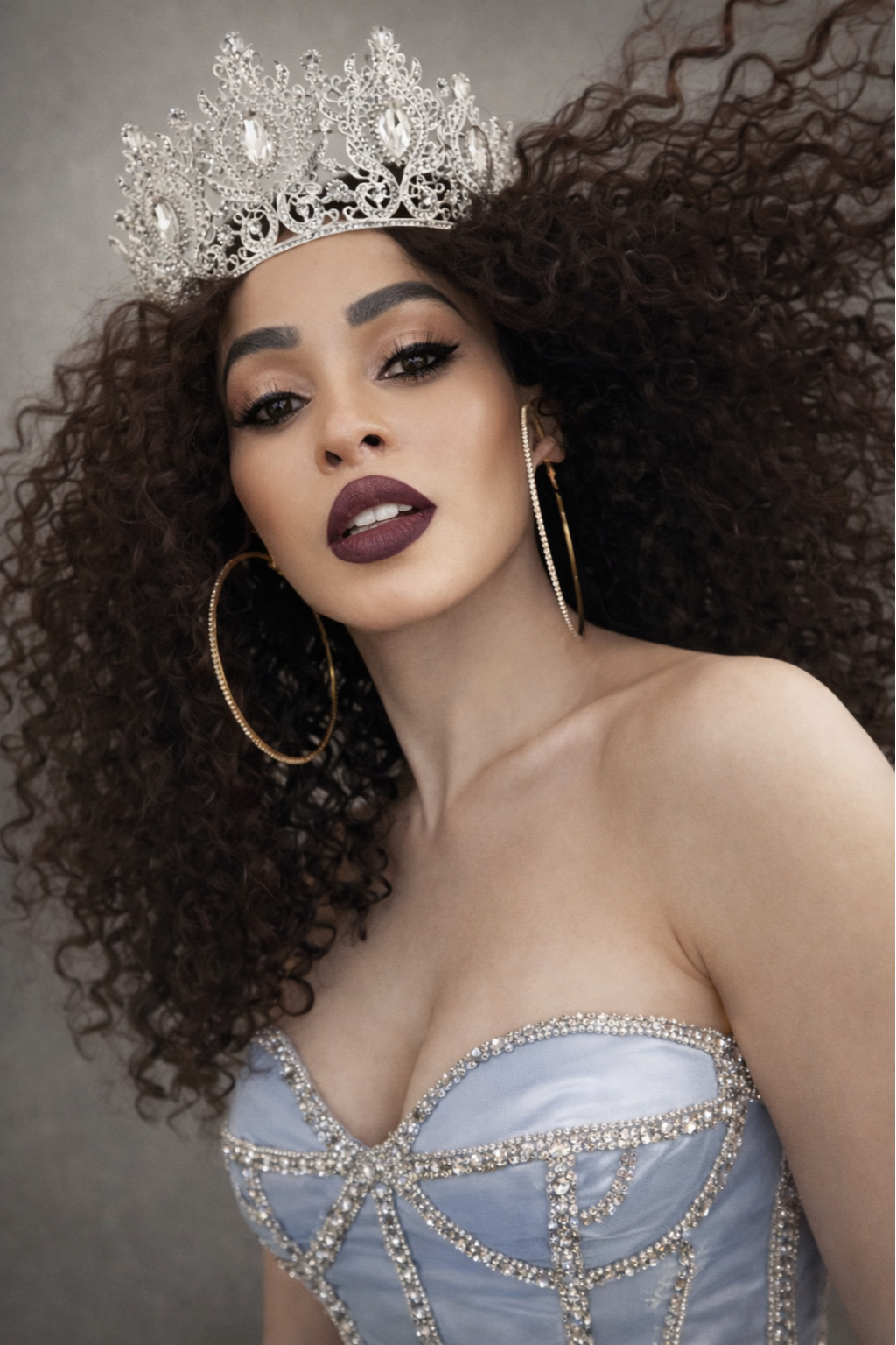 A woman with voluminous curly hair, wearing a large jeweled crown, gold hoop earrings, and an elegant strapless dress decorated with rhinestones.