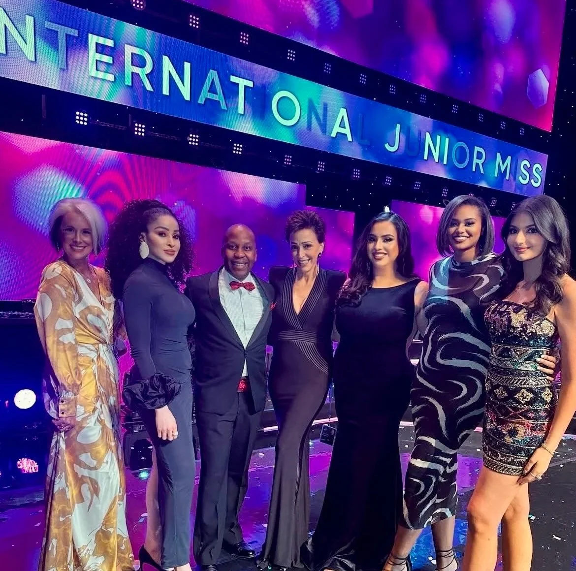Group of women and a man dressed formally on stage during the Miss International Junior pageant, with a vibrant purple and pink background displaying the event name.