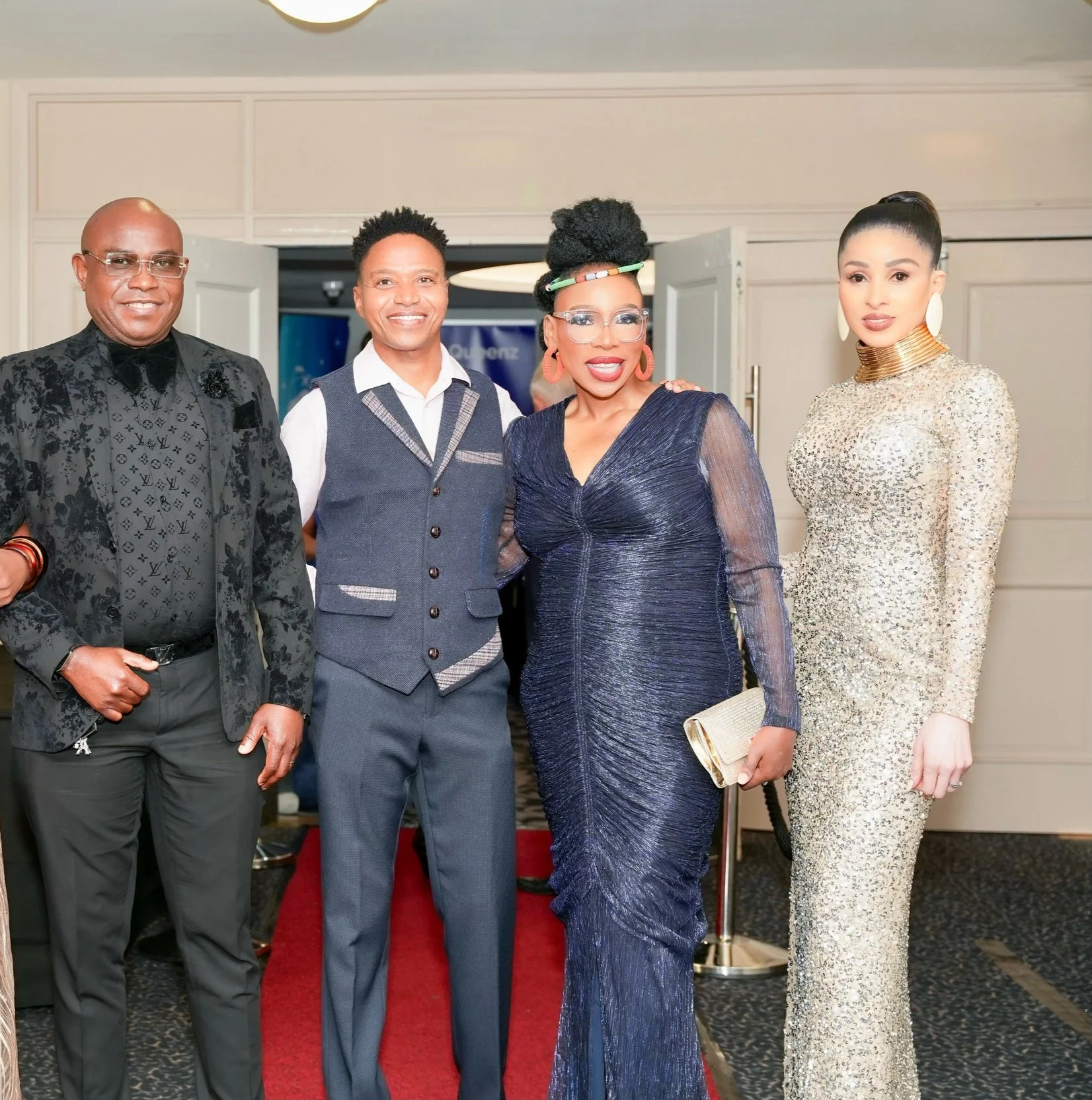 Four people dressed in formal attire posing at an indoor event, with a red carpet, white walls, and doors in the background.
