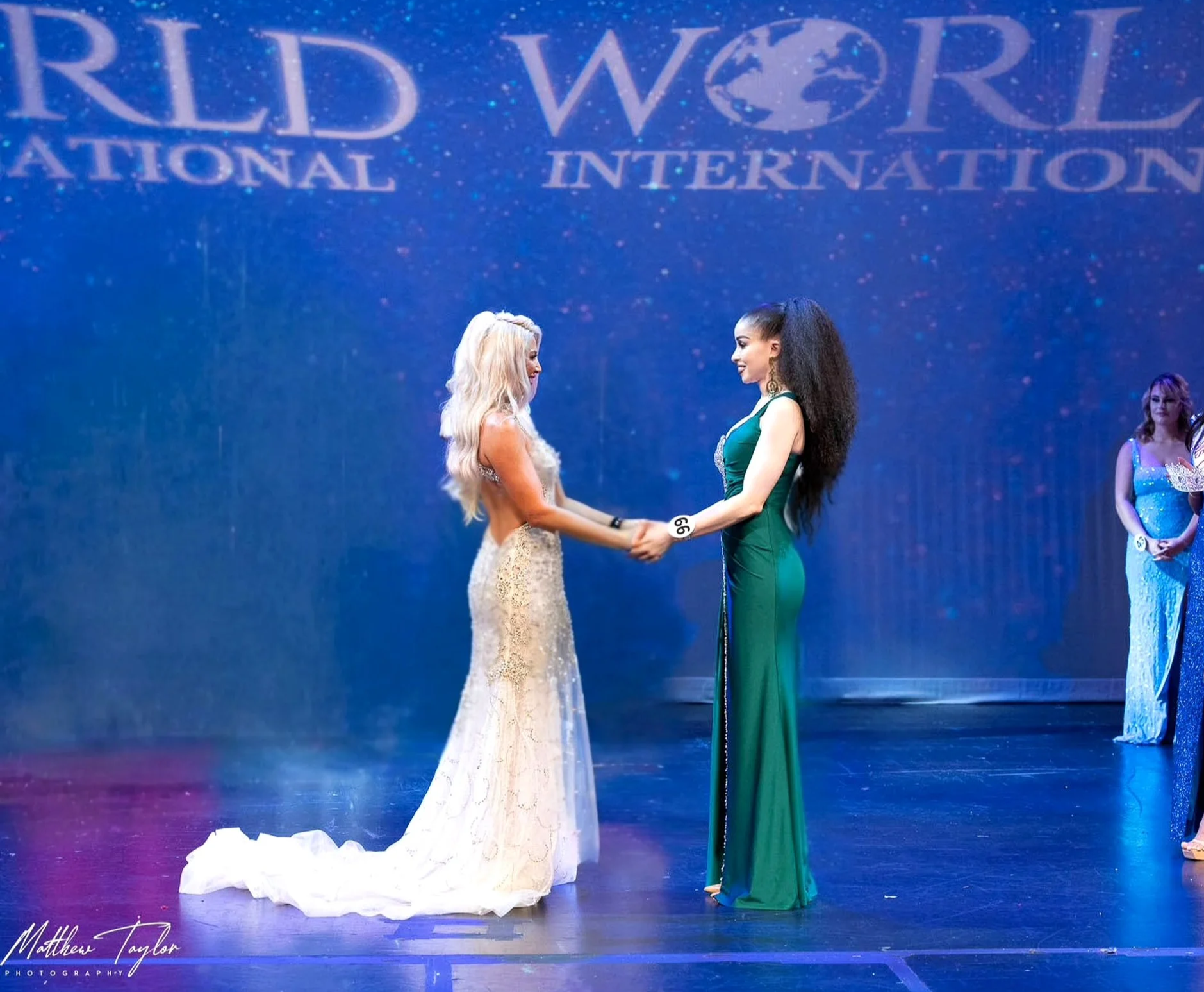 Two women on a stage holding hands during a pageant or competition; one woman in a white dress with blonde hair, the other in a green dress with dark hair. A third woman in a blue dress stands in the background. The stage has a large backdrop with the words 'World International' visible.