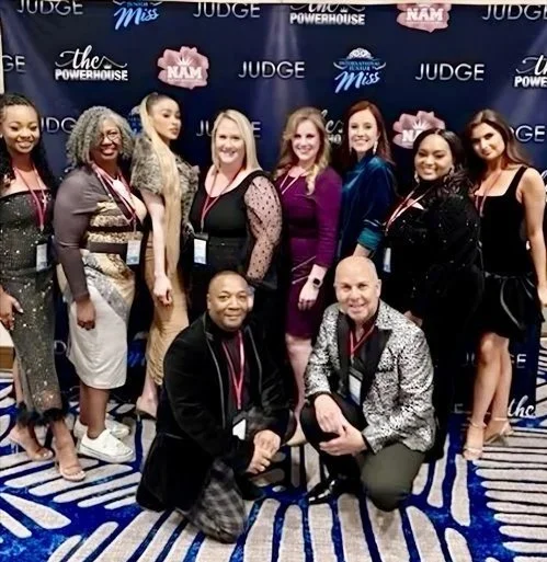 Group of nine women and two men posing together at an event, with a backdrop featuring words like 'Judge,' 'Miss,' and 'Powerhouse.'
