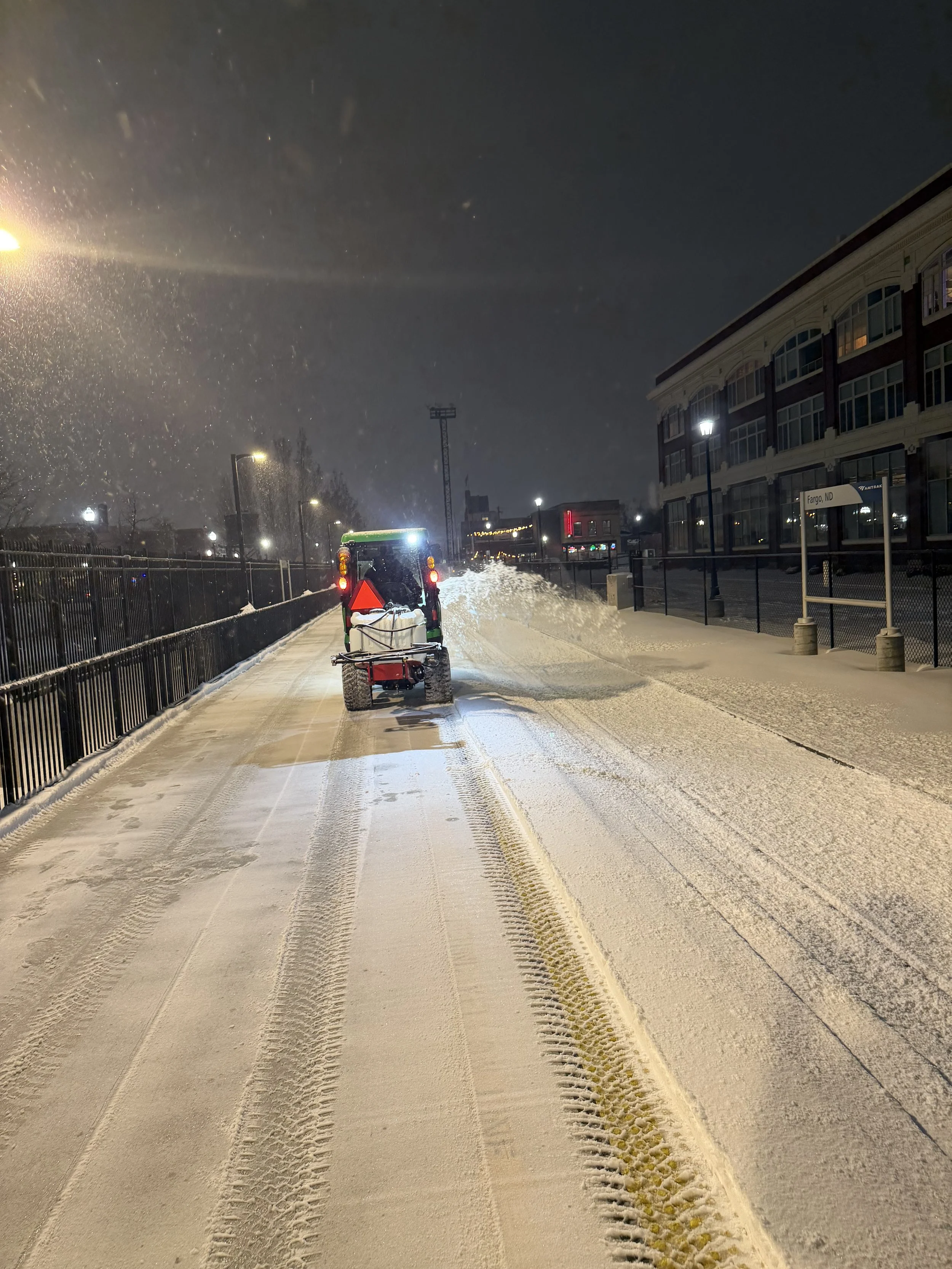 Snow-covered sidewalk and street illuminated by streetlights at night, with a snow plow clearing the path.