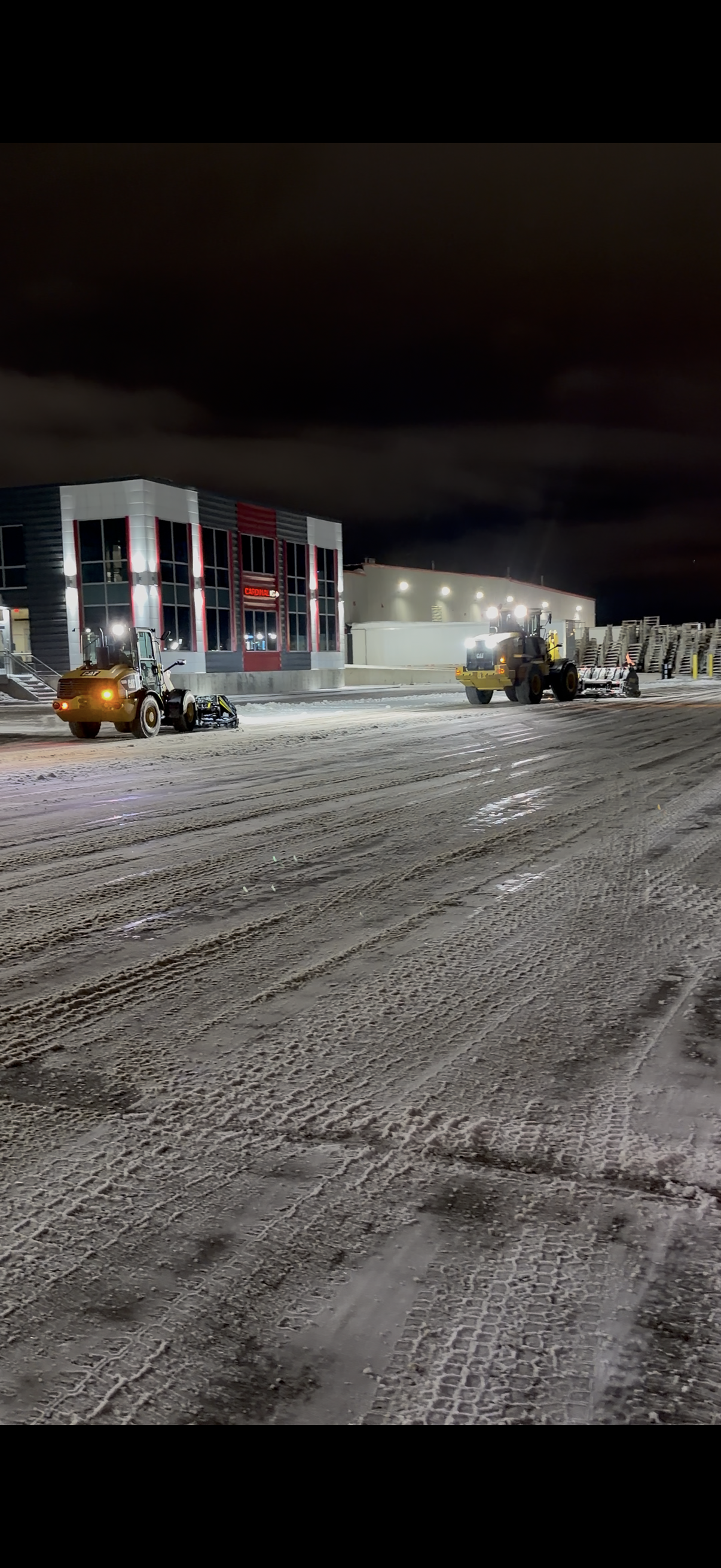 Nighttime scene of a parking lot with snow being cleared by a snow plow, with a modern building in the background.