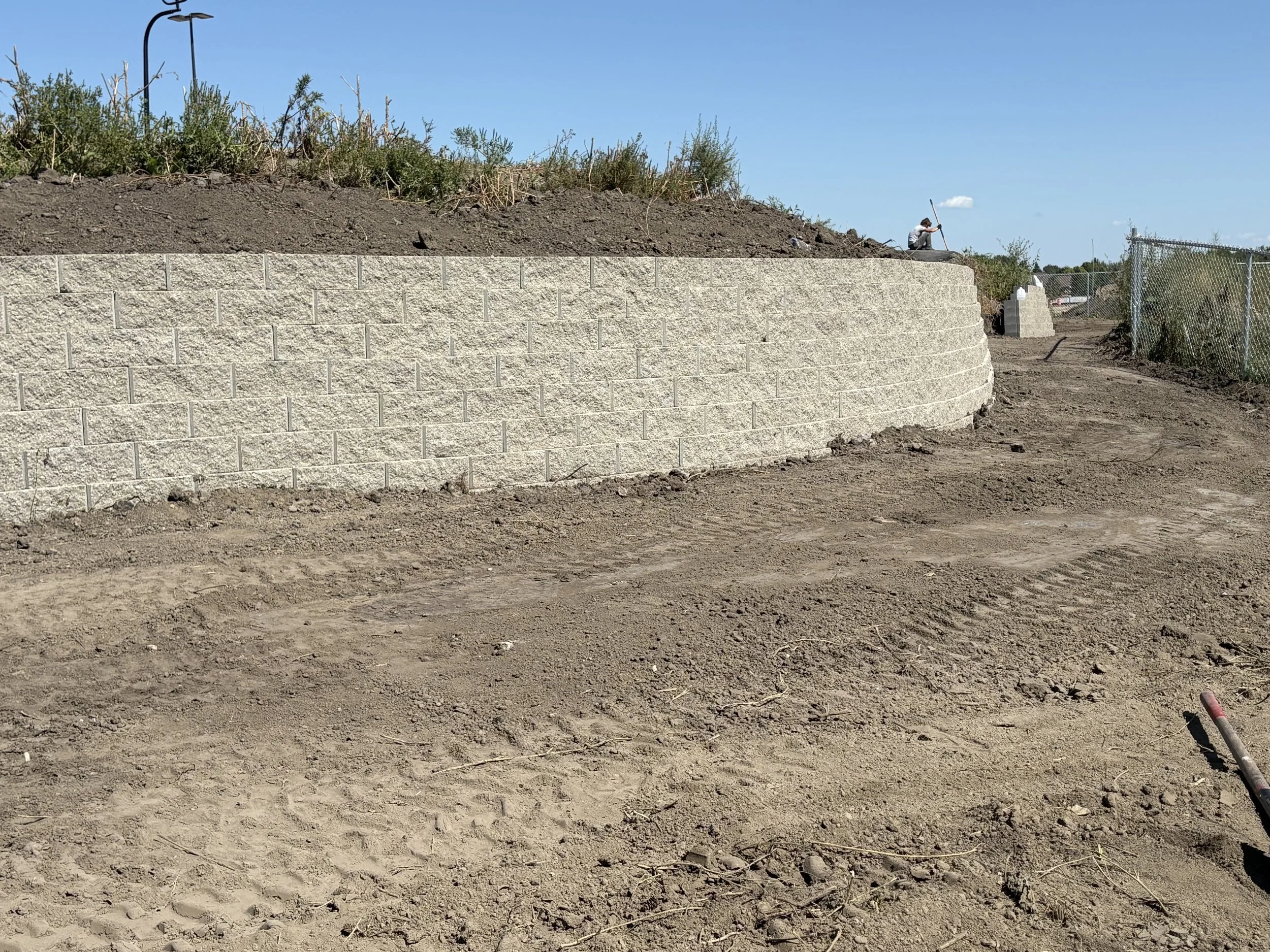 A construction site with a curved retaining wall made of concrete blocks. The ground in front of the wall is dirt, and there is a person working on the top of the wall with a tool in a bright, clear day.