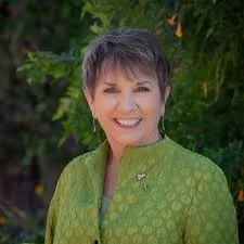 A smiling woman with short gray hair, wearing a green patterned jacket, standing outdoors with greenery in the background.