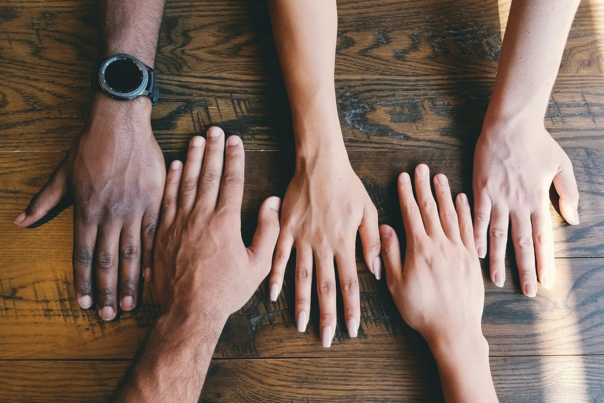 Four hands, two with darker skin and two with lighter skin, are placed on a wooden table, with the hands arranged in a line, palms down.