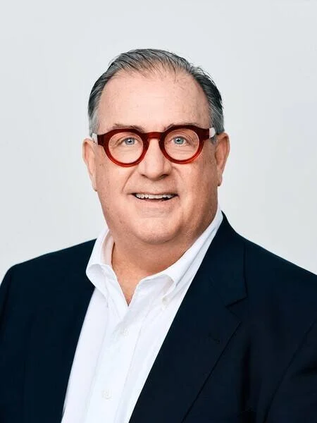 A smiling man with gray hair, wearing a black suit, white shirt, and red round glasses, against a plain light gray background.