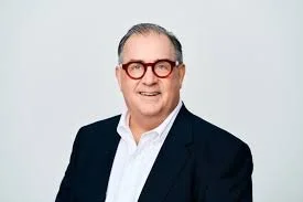 A middle-aged man with short gray hair, wearing glasses, a white shirt, and a dark blazer, smiling against a plain light gray background.