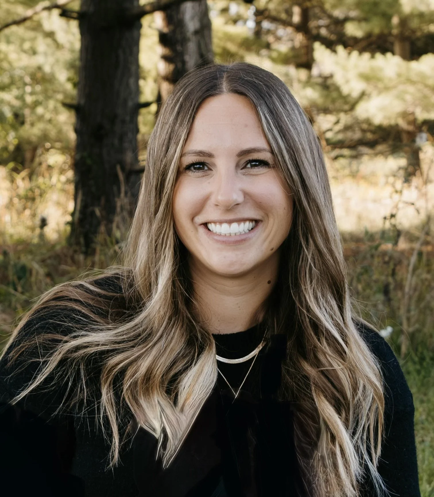 A smiling woman with long wavy hair outdoors, surrounded by trees and green foliage.