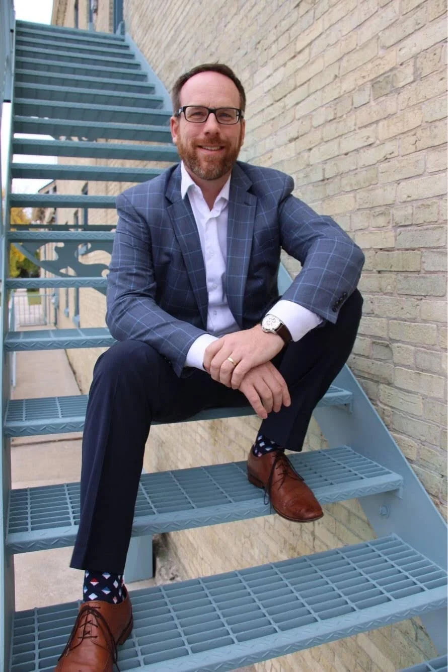 A smiling man with glasses, a beard, and a mustache, sitting on an outdoor metal staircase next to a brick wall. He is wearing a blue plaid suit jacket, white dress shirt, dark pants, brown shoes, and polka dot socks. He is resting his ankle on his knee and has a watch on his left wrist.