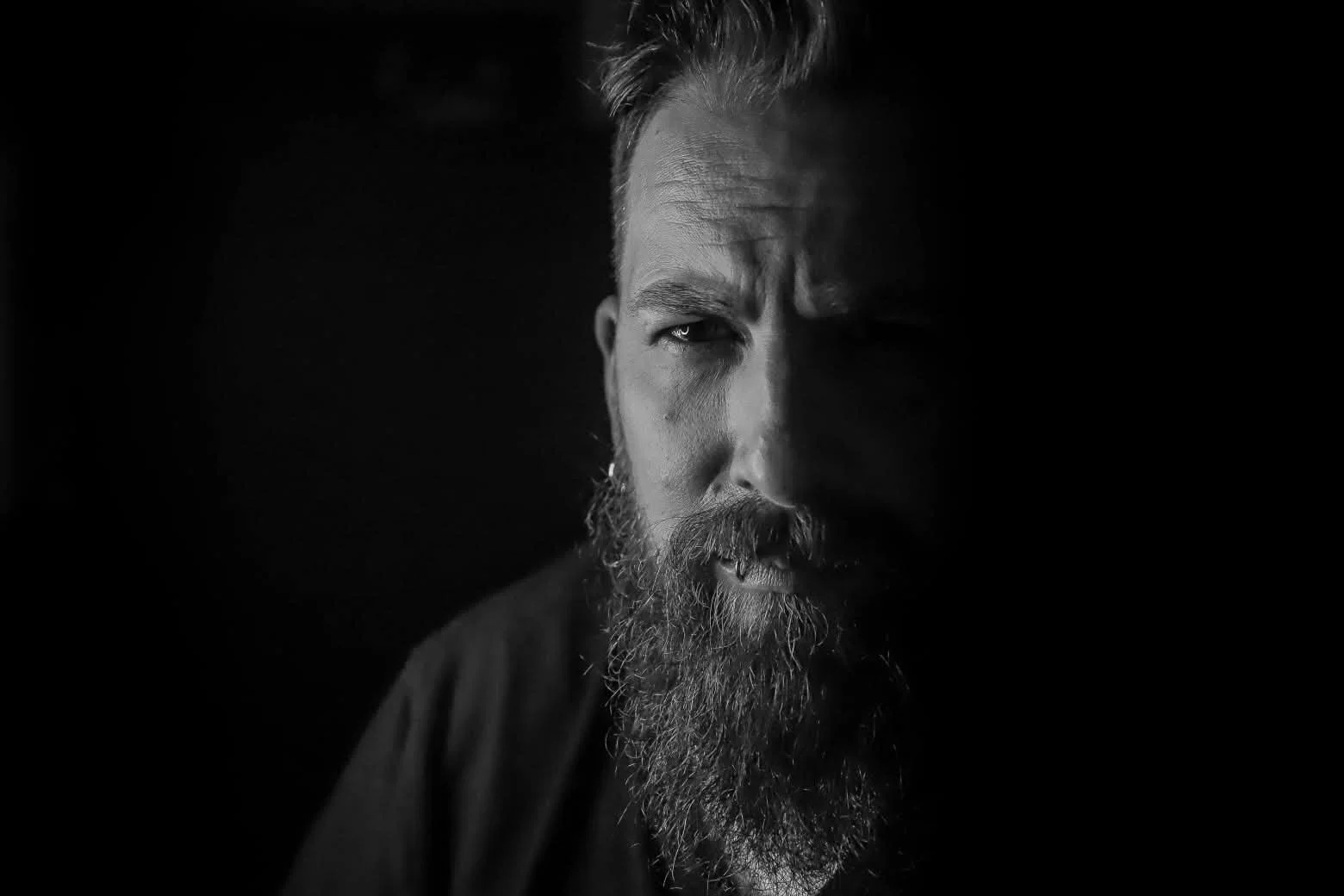 A black and white close-up portrait of a man with a beard, showing half of his face in shadow, staring intensely.