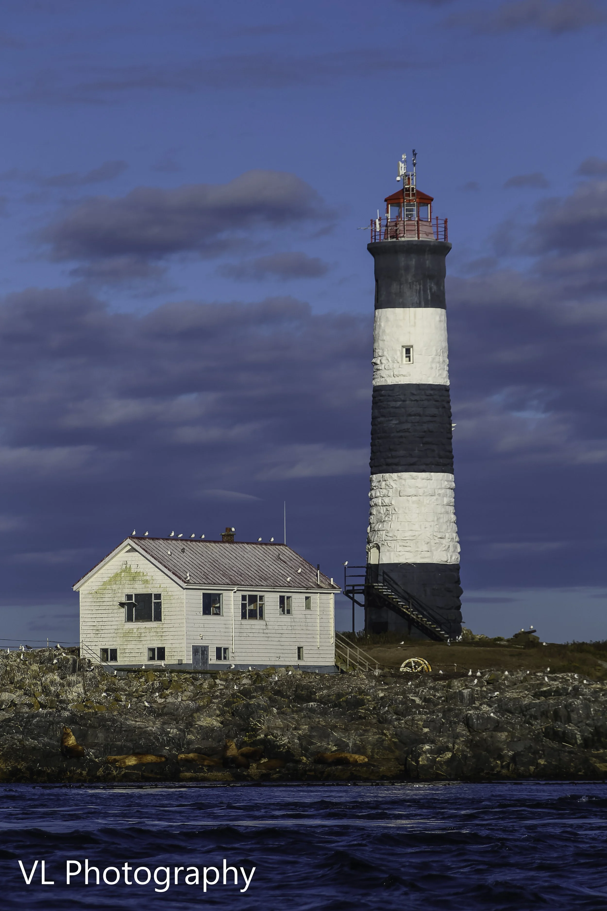 A lighthouse with black and white stripes on a rocky shore, with a small white building nearby and a partly cloudy sky above.