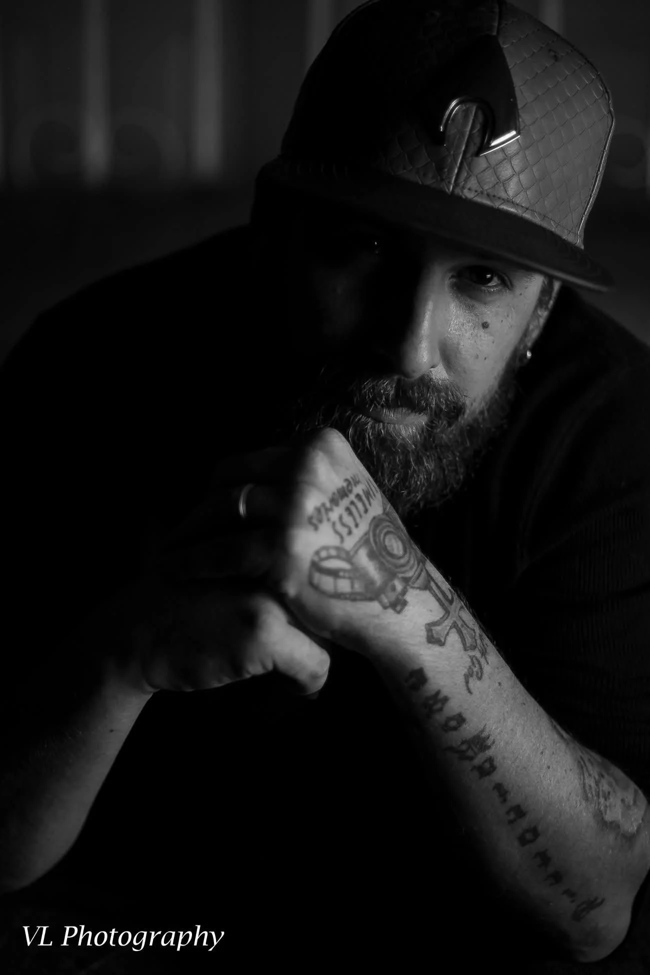 A black and white portrait of a man with a beard wearing a quilted leather cap, resting his chin on his tattooed arm in a thoughtful pose.