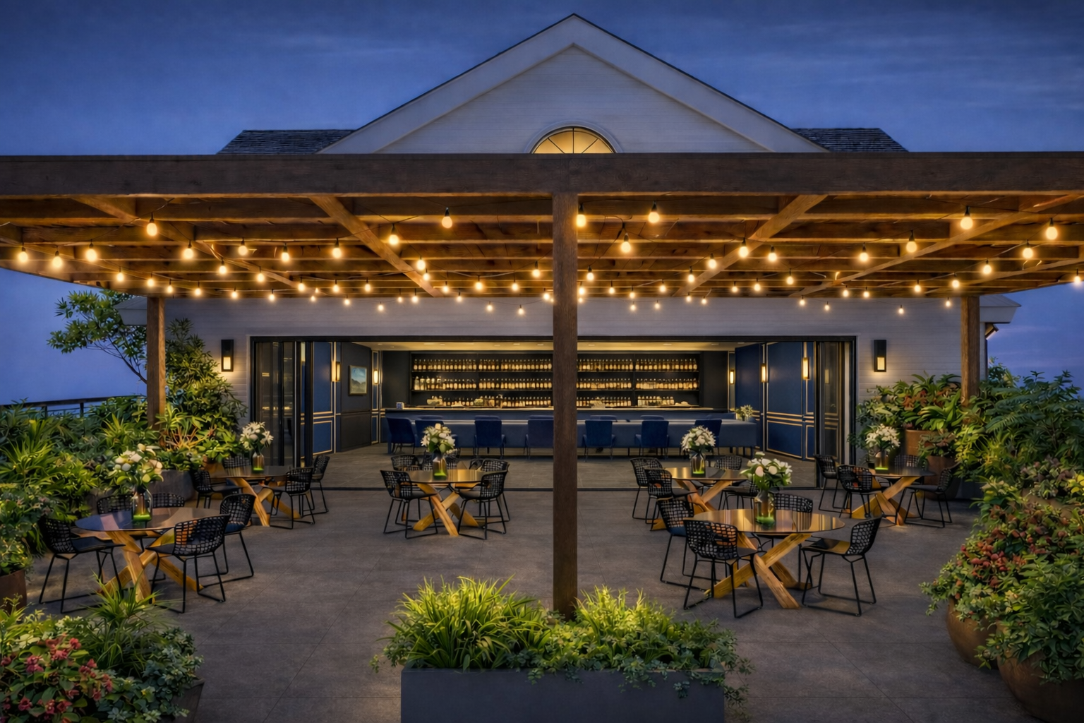 Outdoor rooftop bar and dining area at dusk, with string lights, potted plants, and tables with flowers, overlooking a building with a triangular roof.