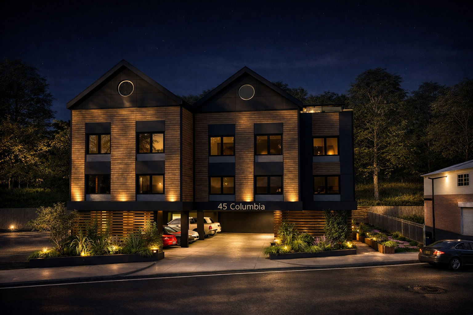 Nighttime view of a modern three-story building with wooden and black exterior, illuminated by warm exterior lights, with cars parked in front and a garden with plants and pathway lighting.