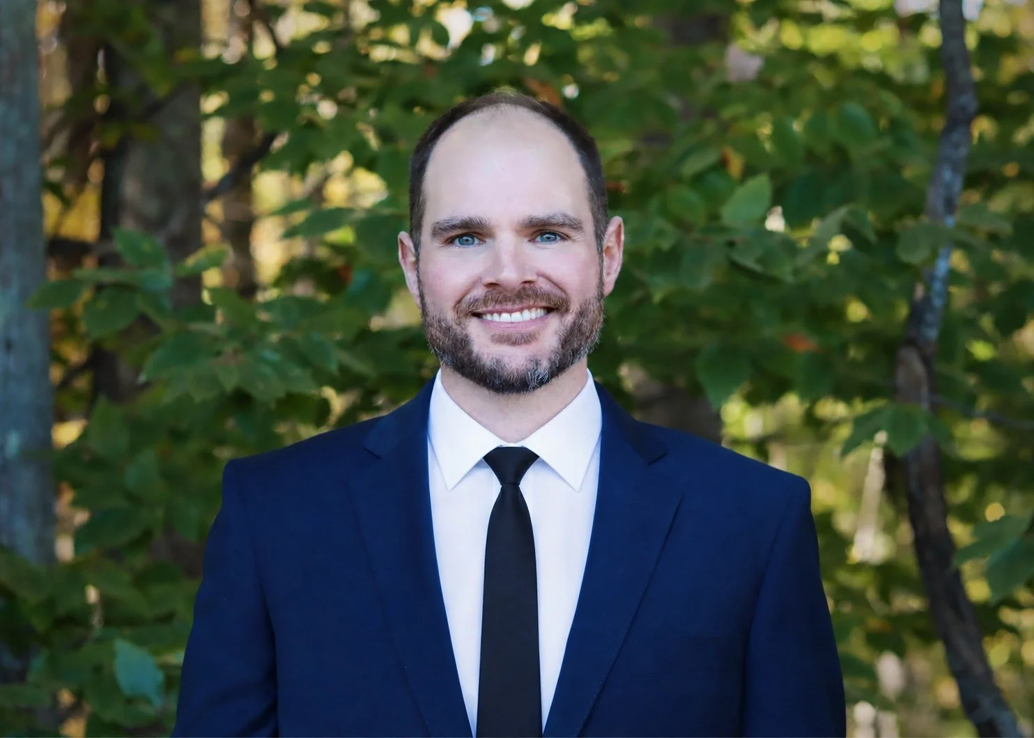 A man with a beard and blue eyes smiling, wearing a dark blue suit, white dress shirt, and black tie, standing outdoors with green foliage in the background.