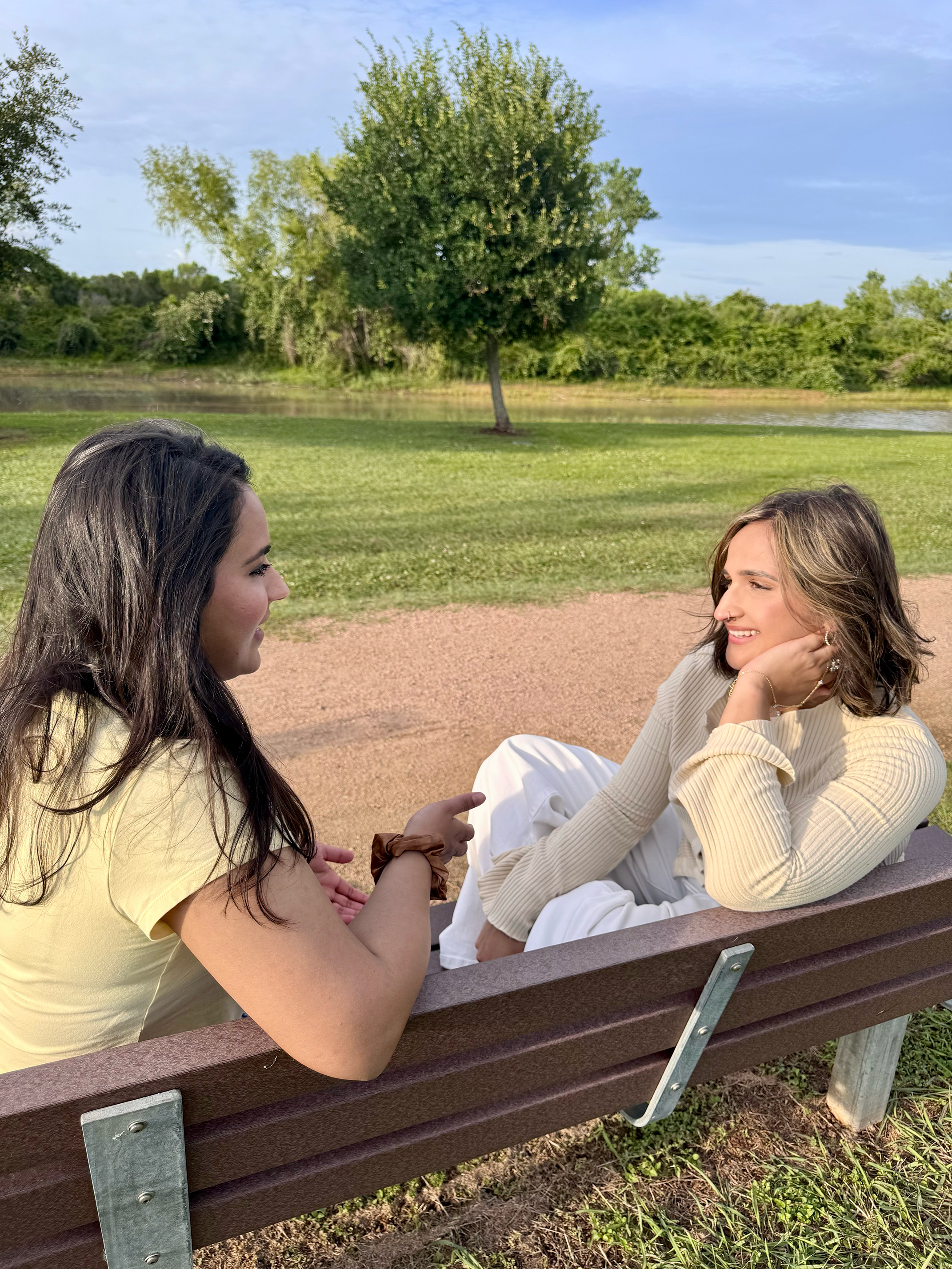 Two women sitting on a park bench facing each other and smiling, with a pond, green grass, trees, and a partly cloudy sky in the background.