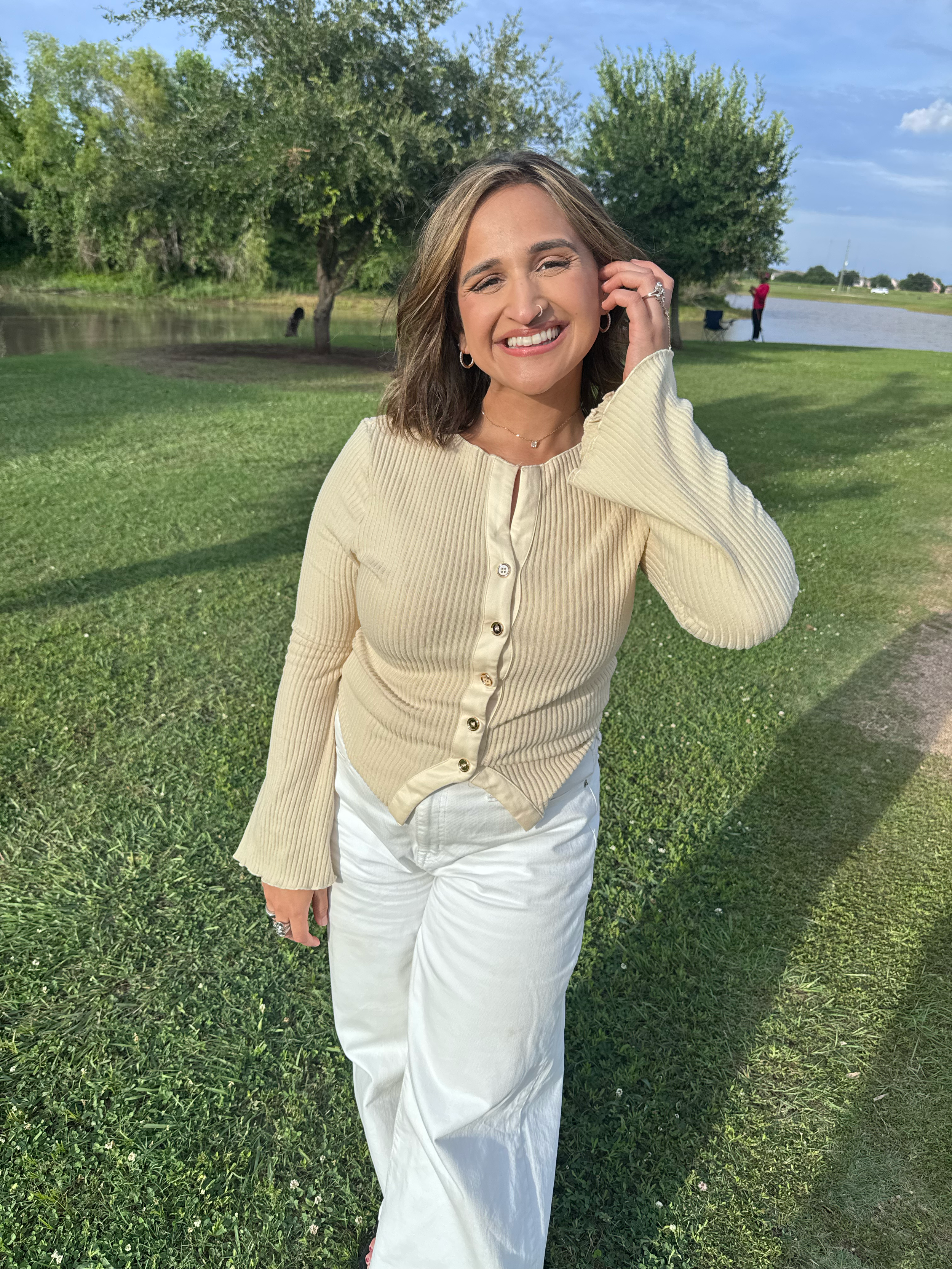 A woman with shoulder-length brown hair, smiling, is standing outdoors on a grassy area near a pond, wearing a light-colored ribbed button-up top and white pants. She is touching her hair with her right hand, and in the background, there are trees and a person fishing near the water.