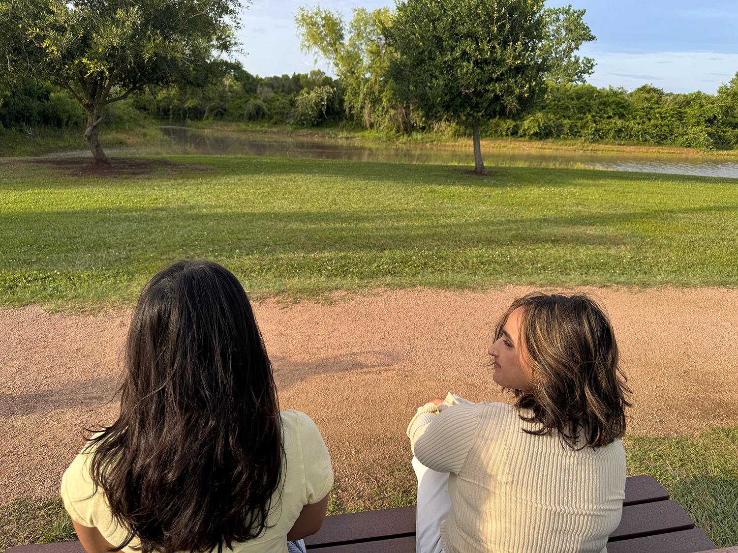 Two women sitting on a bench by a lake, facing away from the camera, with trees and grass in the background on a sunny day.