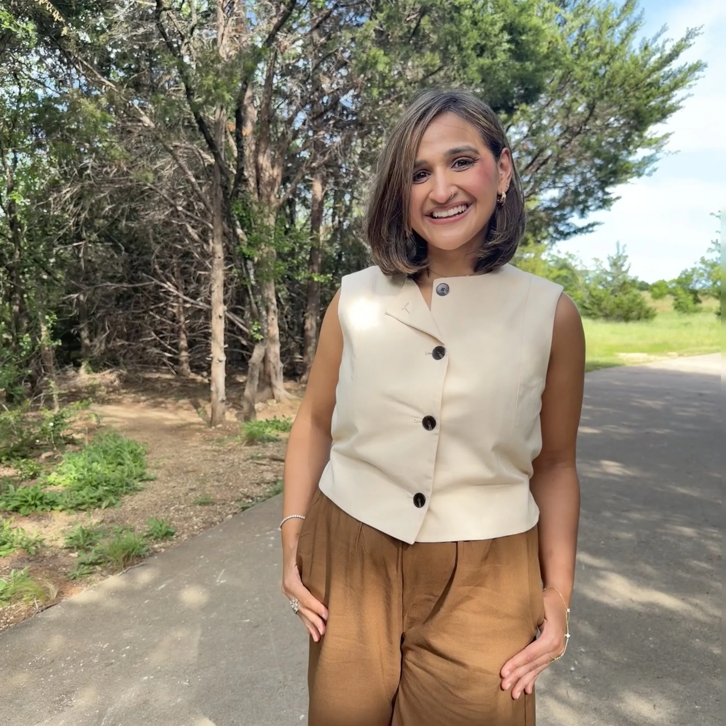 A woman smiling outdoors next to trees, wearing a sleeveless cream-colored top and brown pants.