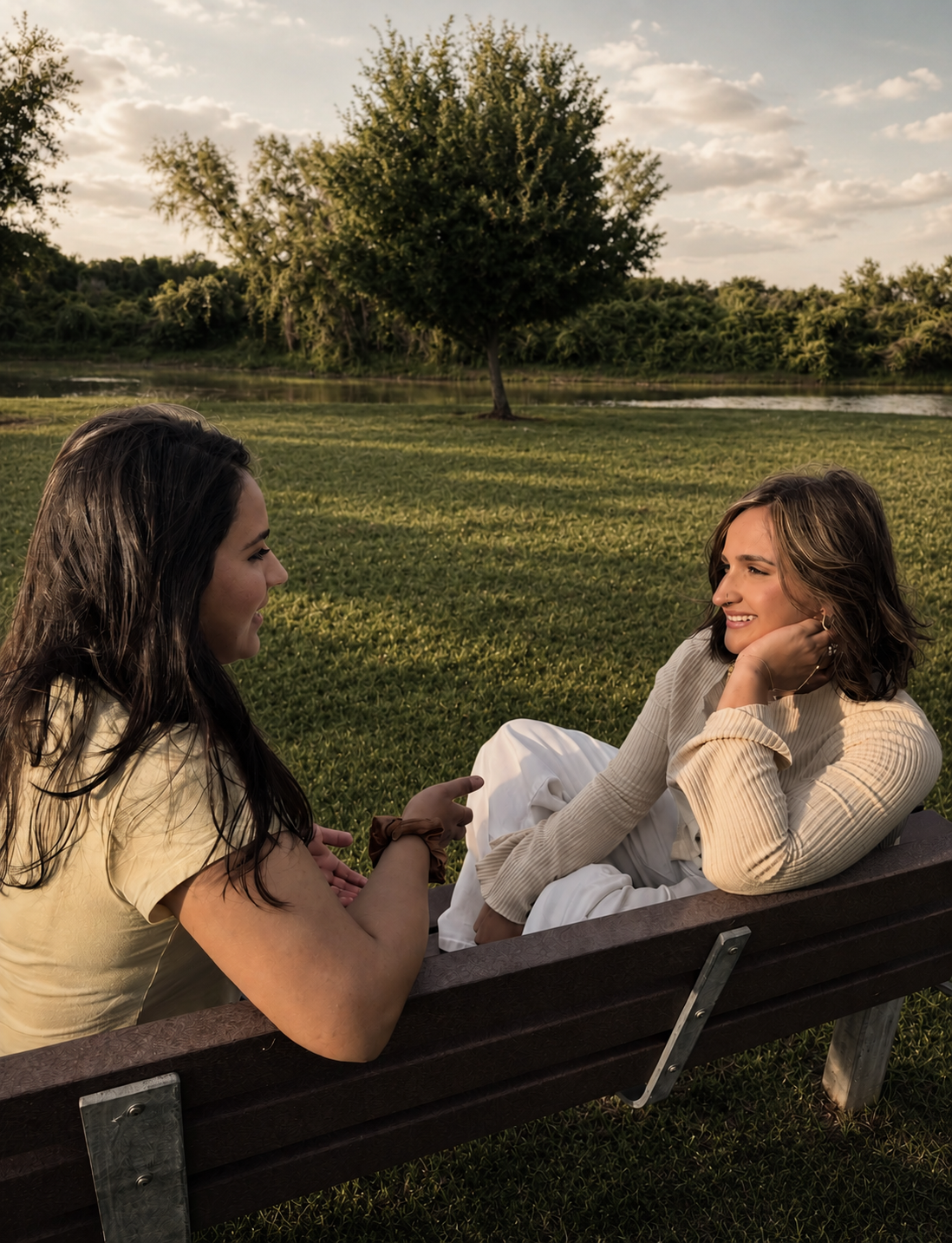 Two women having a conversation on a park bench with a grassy area, trees, and a pond in the background during late afternoon or early evening.
