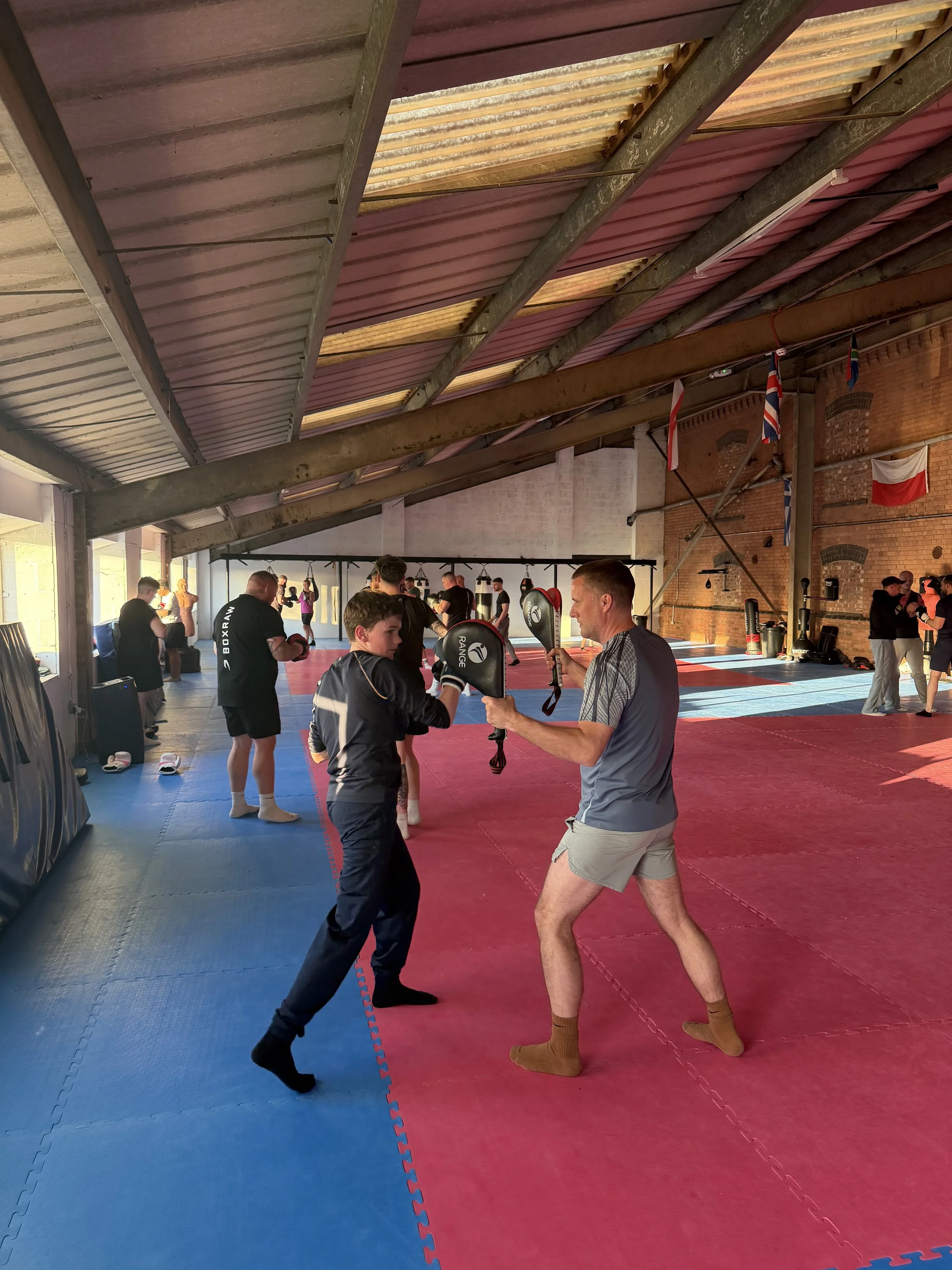 Inside a martial arts gym with people practicing boxing. Two individuals are sparring, with one wearing boxing gloves and pads. Others are standing and observing. The gym has red and blue mats on the floor, flags hanging from the ceiling, and exposed brick walls.