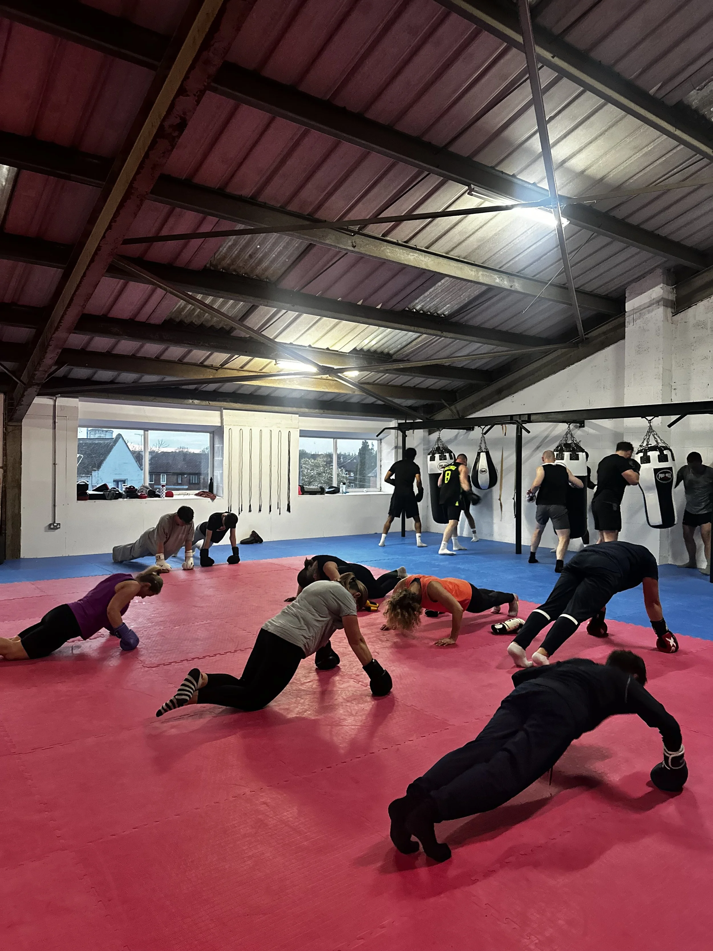 Interior of a boxing gym with a group of people participating in a workout session, doing push-ups or stretching on a red mat, with punching bags hanging on the wall and gym equipment on the side, inside a warehouse-style space with a high ceiling and a window showing a view outside.