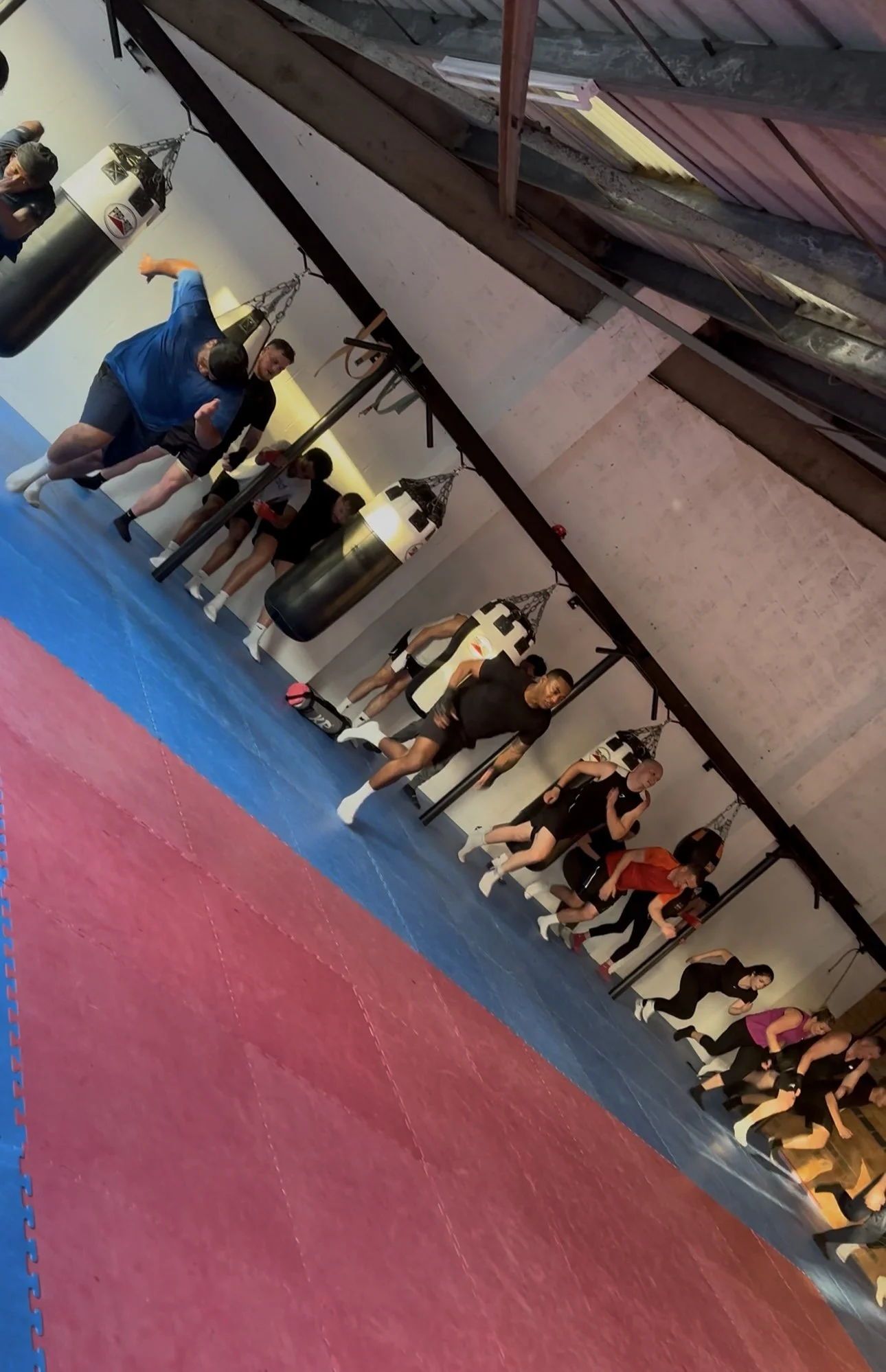 People participating in a martial arts or boxing class, doing stretching and warm-up exercises in a gym with punching bags hanging on the wall.