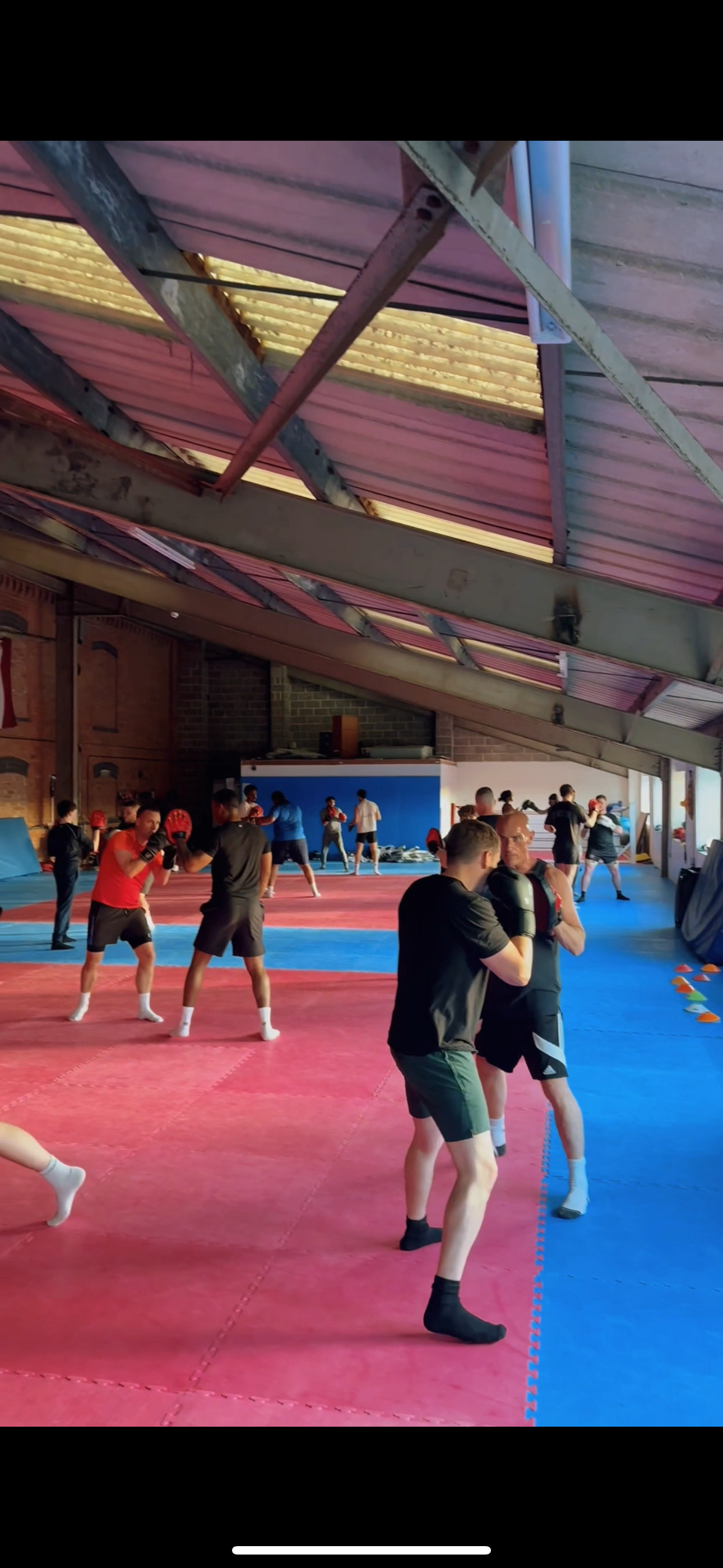 Boxers practicing in a gym with padded flooring, some sparring and others training with focus pads, under a sloped roof with exposed beams.
