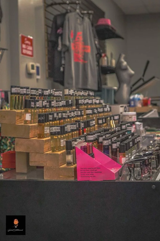 Display of small bottles of perfume or essential oils arranged on a wooden shelf in a store. In the background, there are clothing items, a mannequin, and shelves with other products.
